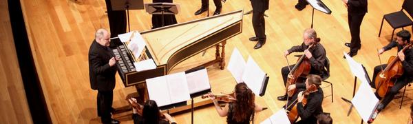 An ensemble of musicians dressed in black stand on stage playing string instruments. The conductor stands in front of them beside a piano.