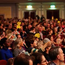 An audience with many children waving paper bees on sticks