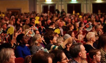 An audience with many children waving paper bees on sticks