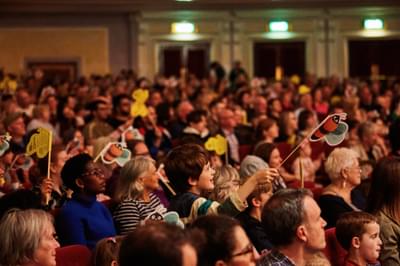 An audience with many children waving paper bees on sticks