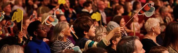 An audience with many children waving paper bees on sticks