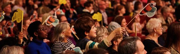 An audience with many children waving paper bees on sticks