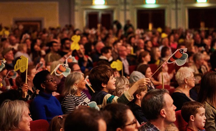 An audience with many children waving paper bees on sticks