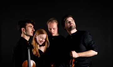 Four musicians stand together all facing different directions. Their black clothing blends into the black background.