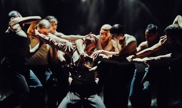 A dancer kneels on the ground with his arms stretched out to the sides. A group of dancers surrounds him reaching out to grab his head, arms and chest.