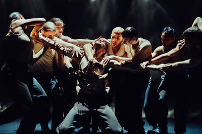 A dancer kneels on the ground with his arms stretched out to the sides. A group of dancers surrounds him reaching out to grab his head, arms and chest.