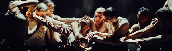 A dancer kneels on the ground with his arms stretched out to the sides. A group of dancers surrounds him reaching out to grab his head, arms and chest.
