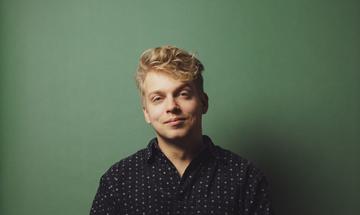 A man sitting facing the camera with one leg crossed over the other. He wears a black spotted shirt and jeans in front of a green background.