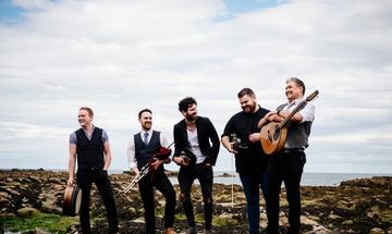 A group of five musicians stand on a rocky landscape with the sea behind them. They are laughing and smiling while holding instruments.