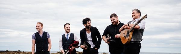 A group of five musicians stand on a rocky landscape with the sea behind them. They are laughing and smiling while holding instruments.