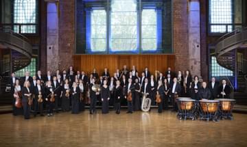 The orchestra stand posed with their instruments in a large room with big windows.