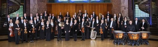 The orchestra stand posed with their instruments in a large room with big windows.