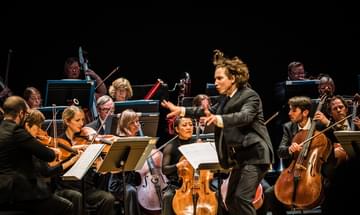 A conductor waves his arms enthusiastically before an orchestra