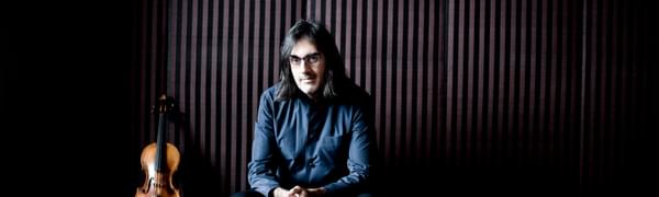 A man with long hair and glasses sits with his hands clasped looking at the viewer. A violin is propped up next to him.