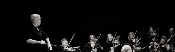 A black and white image of an orchestra sitting on stage. The conductor stands before them pointing at the orchestra.