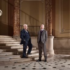 Two men in suits stand looking at the viewer, situated in a grand hall with marble columns