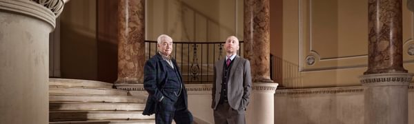 Two men in suits stand looking at the viewer, situated in a grand hall with marble columns