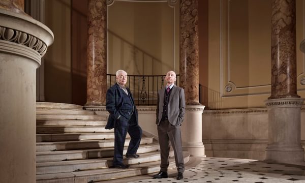 Two men in suits stand looking at the viewer, situated in a grand hall with marble columns