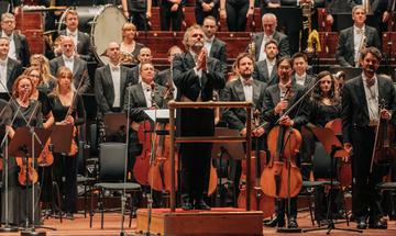 a conductor stands on the platform facing the audience his hands together in gratitude
