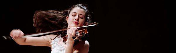 A woman dressed in a white dress playing the violin. Her brown hair is flowing behind her.