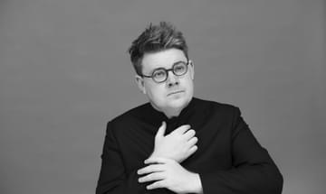 A man with round glasses with both hands held in front of his chest, in black and white