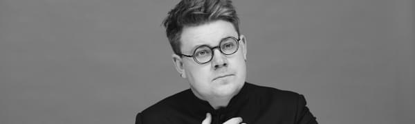 A man with round glasses with both hands held in front of his chest, in black and white