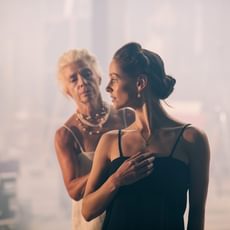 A young woman in black looks over her shoulder. Behind her an older woman in white reaches for her.