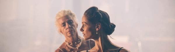 A young woman in black looks over her shoulder. Behind her an older woman in white reaches for her.