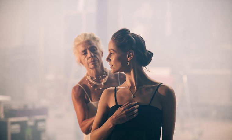 A young woman in black looks over her shoulder. Behind her an older woman in white reaches for her.