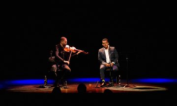 Two musicians wearing suits sit on a dark stage, one playing a violin.