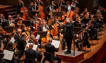 An orchestra appears on stage with the conductor stood before them
