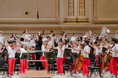 An orchestra of young musicians play their instruments on stage dressed in white tops and red trousers. The conductor stands before them with his hand up in the air.