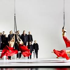 Four dancers perform in red flowing skirts, three of which are suspended from ropes. In the background, a choir dressed in black jumpsuits sing on top of white platforms.
