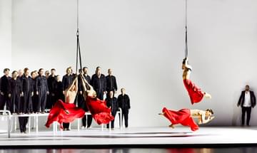 Four dancers perform in red flowing skirts, three of which are suspended from ropes. In the background, a choir dressed in black jumpsuits sing on top of white platforms.