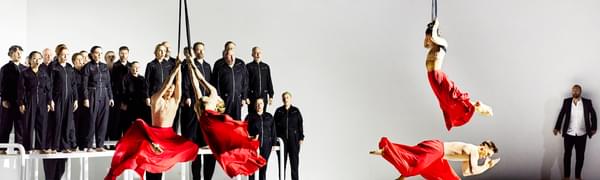 Four dancers perform in red flowing skirts, three of which are suspended from ropes. In the background, a choir dressed in black jumpsuits sing on top of white platforms.