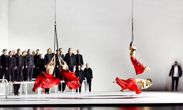 Four dancers perform in red flowing skirts, three of which are suspended from ropes. In the background, a choir dressed in black jumpsuits sing on top of white platforms.