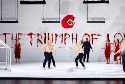 Two shirtless men stand below a woman in a red dress as she spins in the air.