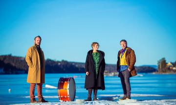 Three musicians stand on a snowy landscape with a bright blue sky. They are dressed in bright colours with a drum sitting next to them.