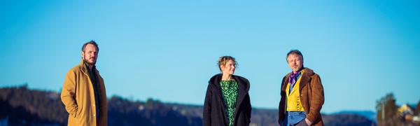 Three musicians stand on a snowy landscape with a bright blue sky. They are dressed in bright colours with a drum sitting next to them.