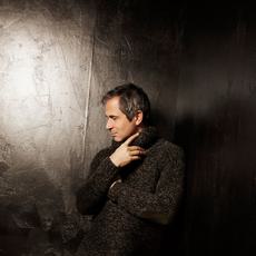 A man stands in a black room facing the side. He is wearing a knitted jumper and black trousers.