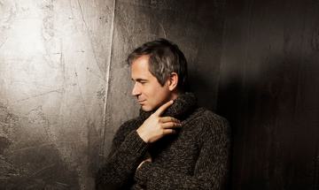 A man stands in a black room facing the side. He is wearing a knitted jumper and black trousers.