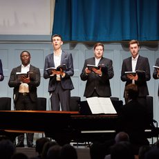 Six men in suits sing on stage holding songbooks.
