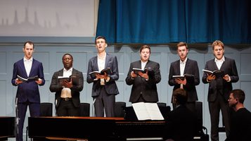 Six men in suits sing on stage holding songbooks.