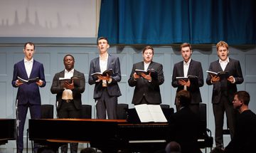 Six men in suits sing on stage holding songbooks.