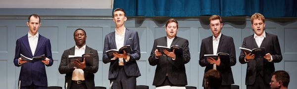 Six men in suits sing on stage holding songbooks.