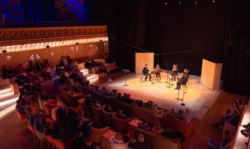 Five musicians playing on a warmly lit stage to an audience.
