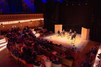 Five musicians playing on a warmly lit stage to an audience.