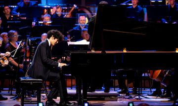 A young man with black hair is playing a grand piano in front of an orchestra