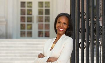 a women stands smiling with her arms folded. she is leaning against a metal gate taller than her and is wearing a white blazer