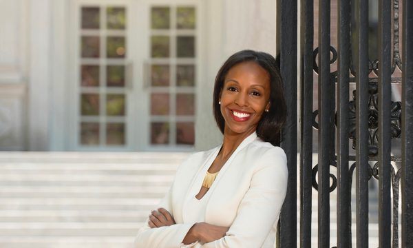 a women stands smiling with her arms folded. she is leaning against a metal gate taller than her and is wearing a white blazer